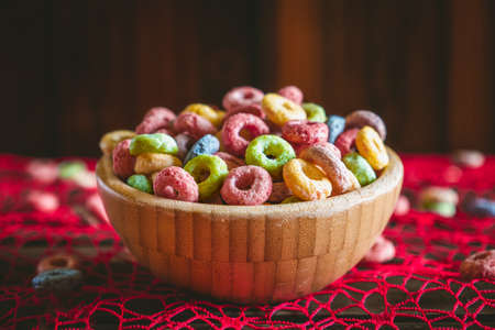 Colorful round fruit cereals in a wooden bowl on a red table, with some scattered outside the bowl. Wooden background behind with copy space. Breakfast ready.の写真素材