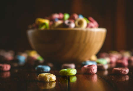 Closeup colorful round fruit cereals scattered on a dark wooden table with a bowl full of cereals behind.の写真素材