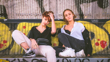 Two girls sitting on a bench against a wall full of graffiti on the street.の写真素材