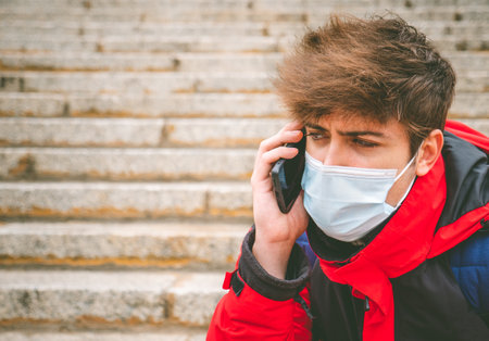 Angry guy with face mask and backpack talking on the phone in street stairs on a winter day.の写真素材