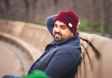 Boarded guy in a woolen hat and sweater sitting on park bench. Looking at the camera and smiling.の写真素材