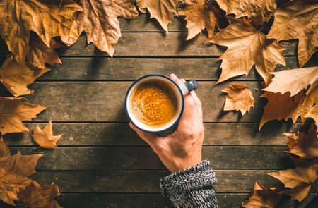 Hand of a caucasian male holding a hot coffee cup on an aged wooden table with autumn leaves around it. Homey feeling and relax.の写真素材