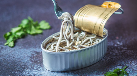 Fork pricking canned baby eels on a blue table with parsley.