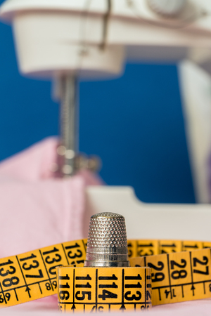 Thimble on yellow tape measure, and white sewing machine. Pink and blue background. Sewing hobby concept. Verticalの写真素材