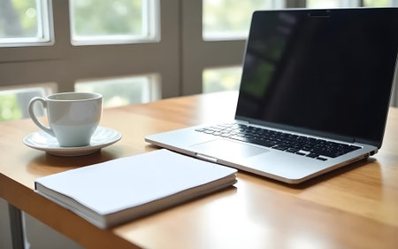 A wooden desk by a window, featuring a laptop, a disposable coffee cup, a notebook, and a pen,...