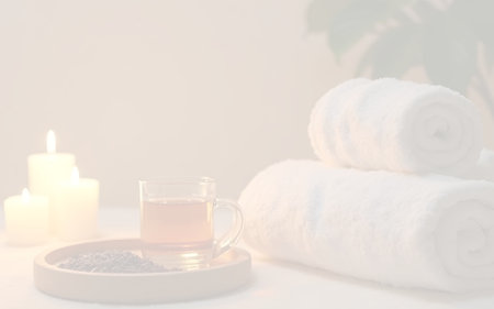 A serene and warm ambiance captured, depicting a moment of relaxation with three softly glowing white candles. An amber-colored herbal tea in a glass mug sits on a wooden tray with what appears to be coffee beans or dried herbs, alongside neatly rolled clean white towels. A green plant in the background adds a touch of freshness, perfect for conveying self-care, wellness, and spa themes.の写真素材