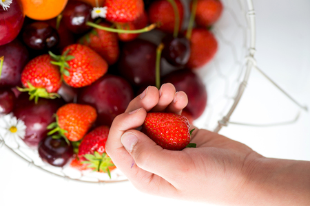 Hand holding Strawberry above Summer Berries and Fruits in the basketの写真素材