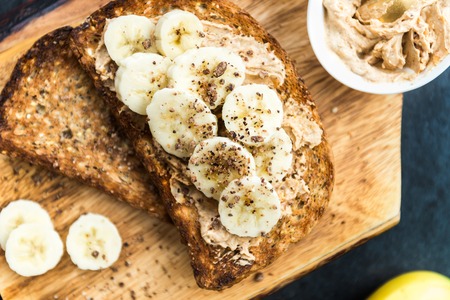 Toasts from Wholewheat Seeded Bread with Peanut Butter and Banana on the Wooden Board on dark marble...