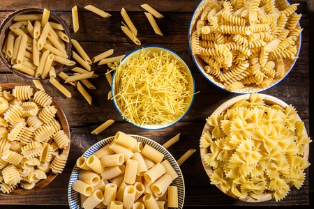 Different Types of Raw Pastas such as Fusilli, Penne Rigate, Radiatori, Farfalle, Mezzi Rigatoni and Egg Noodles in Bowls on Wooden Rustic Backgroundの写真素材