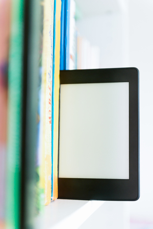 Ebook or digital reading tablet device standing on the shelf with bright real books の写真素材