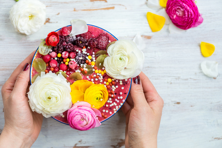 Hands are holding Bowl with Breakfast Detox Pink Smoothie from Banana and Forest Berries, topped with frozen berries and ranunculus flowers, light wooden background, top viewの写真素材