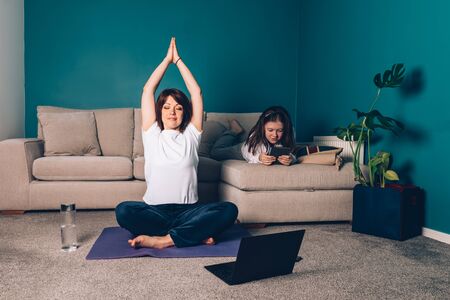 Woman is doing online yoga with laptop during self isolation at her living room, no equipment workout, meditation tips for beginners. Her daughter is reading. Family time with kids, stay home.の写真素材