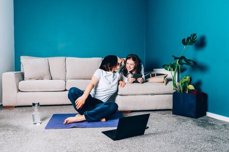 Woman is doing online yoga with laptop during self isolation at her living room, no equipment workout, meditation tips for beginners. Her daughter is reading. Family time with kids, stay home.の写真素材