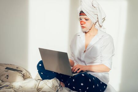 Young woman is working from home and her bed using laptop and phone during self isolation. She is wearing towel on her head and funny face mask. Early morning routine, online work, distance jobの写真素材