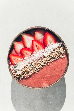 Pink Smoothie from Banana and Strawberries with pieces of Strawberries, Granola and Coconut Shreds on top in Coconut Shell Bowl, strong shadows. Healthy summer breakfast, top viewの写真素材