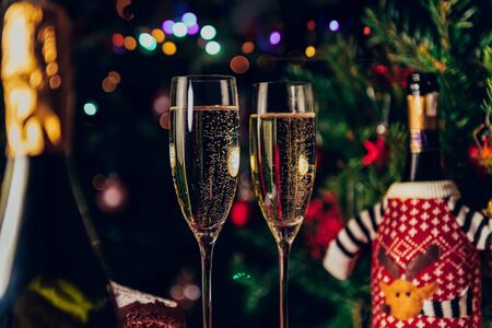 Two flute glasses and bottles of champagne in preparation for Xmas or New Year party. One bottle is in the christmas jumper and xmas tree on the backgroundの写真素材