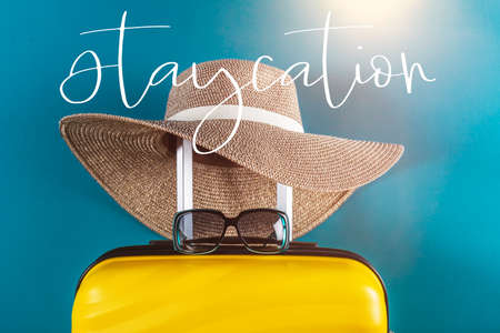 Staycation word. Bright and stylish cabin size suitcase with straw hat against bright blue background. Easy travel at home country during coronavirus outbreak with little baggage concept.の写真素材