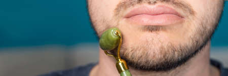 Bearded man is using jade face roller for beauty facial massage therapy, banner sizeの写真素材