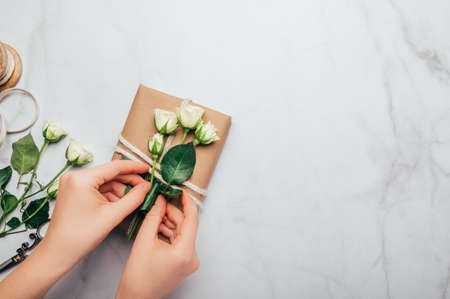 Womans hands are wrapping gifts in recycled brown paper and decorating with real fresh roses, on...