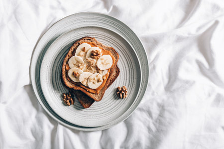 Open sandwich from slice of wholegrain bread with peanut nut butter, bananas and crushed walnuts. Breakfast in bed with plate on beddingの写真素材