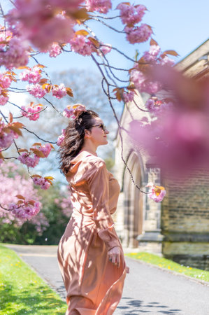 Woman in her mid 30 in park with blooming cherry tree or sakura, springtime in UK. Enjoying life and sunny day with warm weatherの写真素材