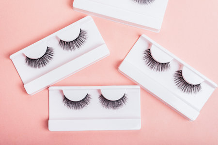 Different fake eyelashes in boxes on trendy pastel pink background. Makeup accessories. cosmetics...