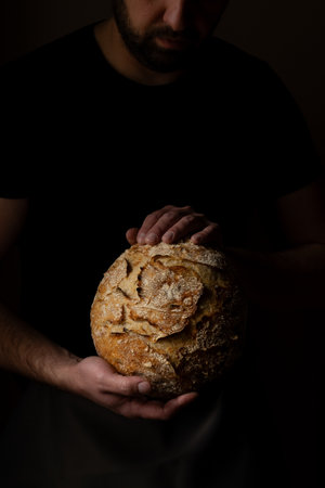 Attractive young Caucasian chef posing with white sourdough bread. The sourdough bread is the central protagonist of the scene, standing out with beautiful golden tones against the dark background.の写真素材