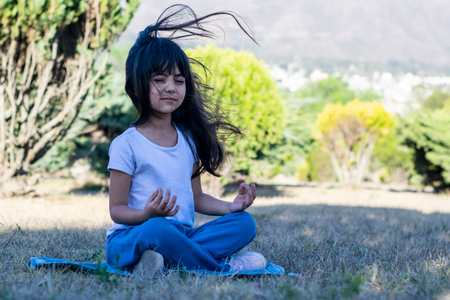 Latin kid outdoors practicing yoga in a park during the day with her hair blowing in the wind.の写真素材