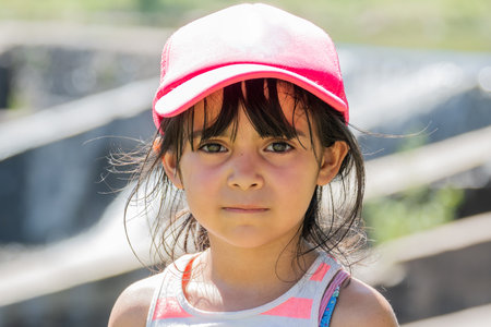 Very close portrait of a latin kid on a trip. The girl wears a pink cap. She looks at the camera with a very serious face.の写真素材