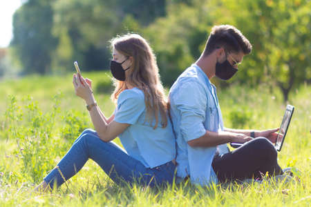 Two young person sitting in the park use laptop and smartphone with face mask