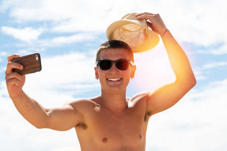 smiling young man with hat and sunglasses taking selfie with smartphone on the beach - smiling teen with martphone taking selfieの写真素材