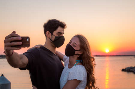 a young couple take a selfie at sunset wearing face mask - selfie during coronavirus - concept about new normalの写真素材