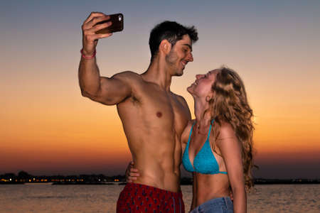 young couple take selfie in the sunset light - two smiling young lovers take selfie on the beach in...