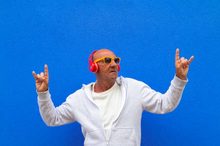 senior man listen music and have fun making rock gesture on blue background - portrait on colored...