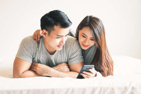 A young Asian couple plays on the phone in bed in the morning in the bedroom. Happy family conceptの写真素材