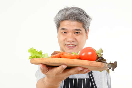 Fat old man holds a wooden tray with fresh vegetables for water to cook. The concept of healthy eating for the elderly. White backgroundの写真素材