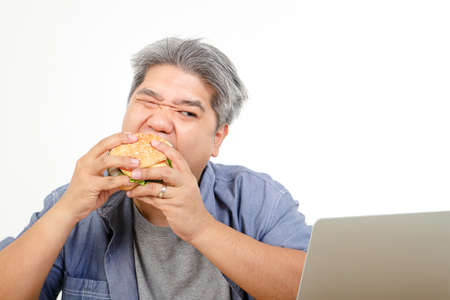 Fat old man eating a hamburger and sitting to work. The concept of healthy eating for the elderly. White backgroundの写真素材