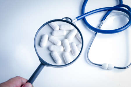 The doctor holds a magnifying glass to see the white pill to treat the common cold and the virus. The concept of careful medication according to doctor's adviceの写真素材