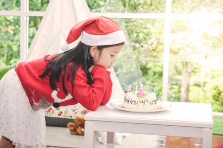 A little girl wearing a red santa claus hat Blow candles, cakes, and wishes for Christmas presentsの写真素材