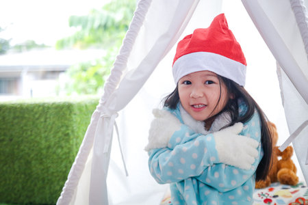 A cute little girl wearing gloves and a red Santa Claus hat sitting in a white tent. Waiting for a gift at Christmasの写真素材