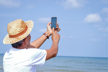 An elderly Asian man holding a phone to take pictures in the seaの写真素材