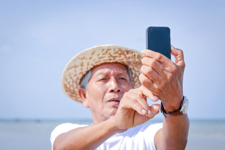 An elderly Asian man holding a phone to take pictures in the seaの写真素材