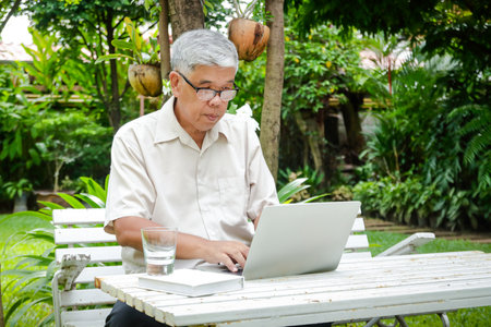 An elderly Asian man sitting at work typing a laptop at the front garden. Retirement and health care conceptsの写真素材