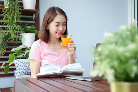 A happy smiling asian woman holding a glass of orange juice And read books Sit back and relax on the balcony. Concept of health problems for women in their 40s. Eat a good and healthy diet.の写真素材