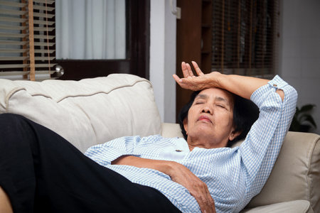 An elderly Asian woman is lying stressed on the sofa at home. Place your hand on your forehead. Senior health care conceptの写真素材