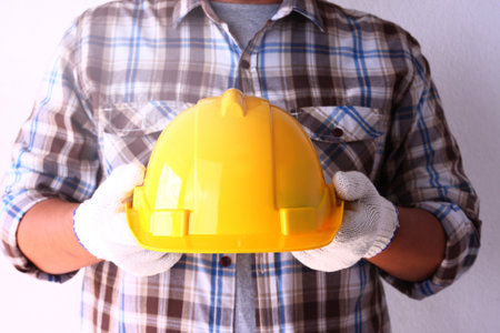 The construction worker wears white gloves, holding a yellow safety helmet for work safety.