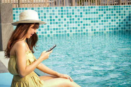 Beautiful Asian woman sitting by pressing the mobile phone at the pool side.