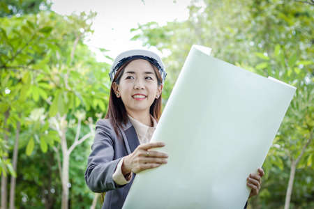 Female environmental engineer wearing a suit, wearing a white hat, standing holding a paper planの写真素材
