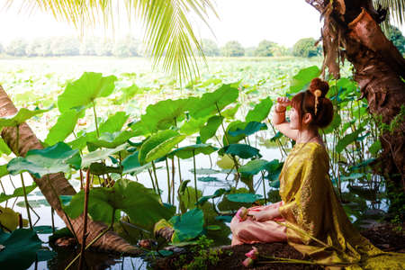 A beautiful woman wearing an ancient Thai dress, sitting at the waterfront in a lotus pond.の写真素材