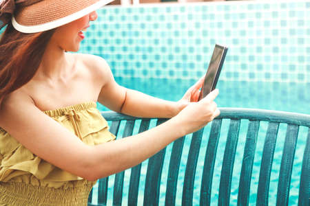A beautiful girl wearing a hat, sitting on a chair, holding a phone, taking pictures at the pool side on holidayの写真素材